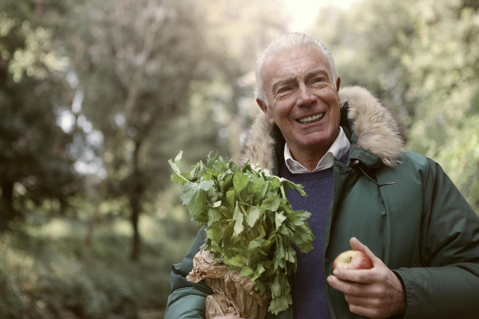 Smiling senior man holds fresh vegetables and an apple in a sunny outdoor setting.